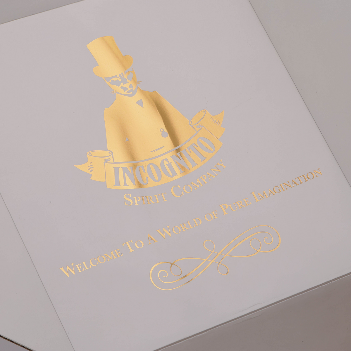 A close-up of a white box lid featuring elegant gold foil branding for 'Incoonto Spirit Company' with a stylized figure in a top hat and the text 'Welcome To A World of Pure Imagination'.