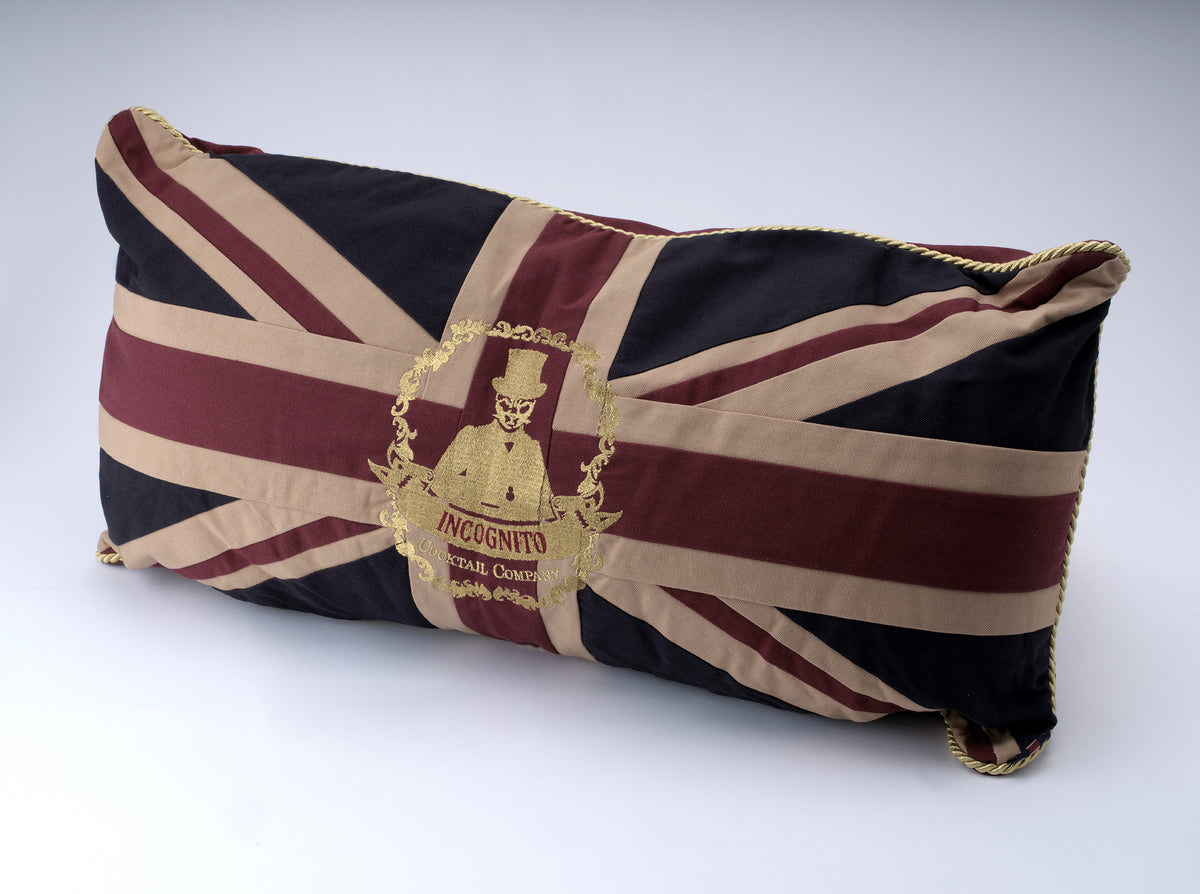 Incognito Union Jack cushion in burgundy, navy and neutral tones with gold rope piping and embroidered Incognito Cocktail Company emblem.