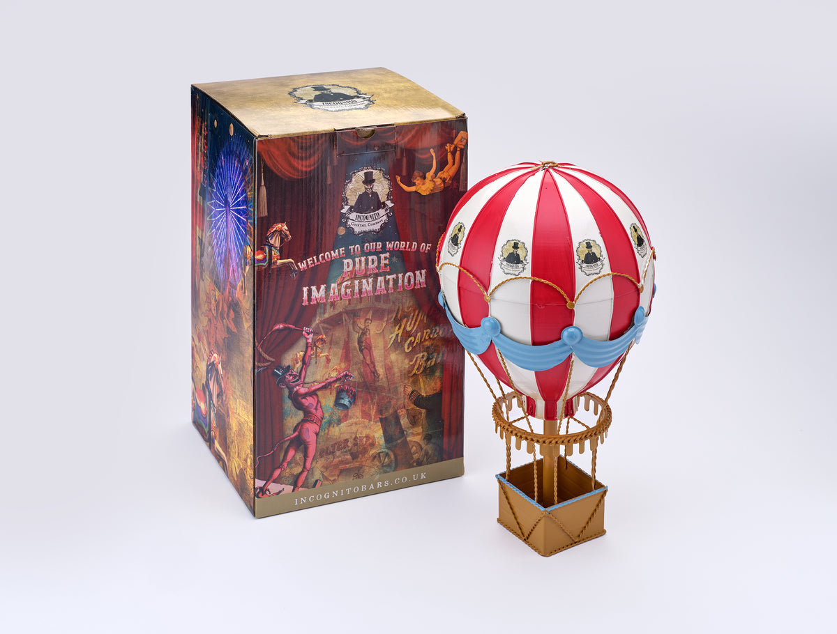 Incognito Hot Air Balloon ornament with red and white striped balloon, blue draping and gold detailing, presented with an illustrated Incognito gift box.